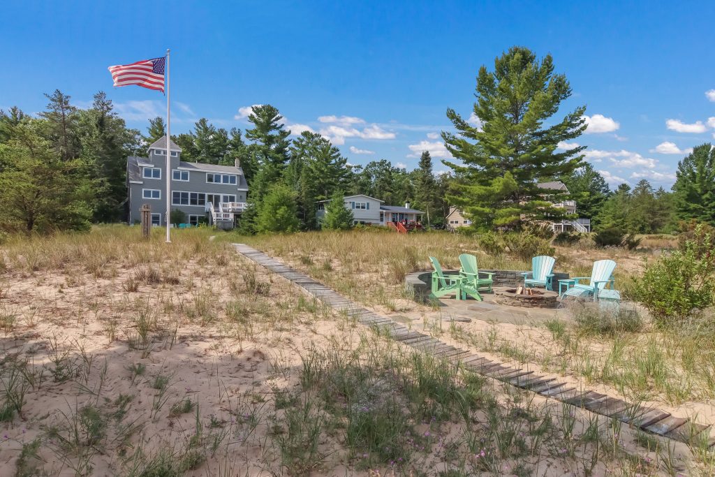 Sandy dune with beach grass and teal chairs beneath a flagpole and pines near the house