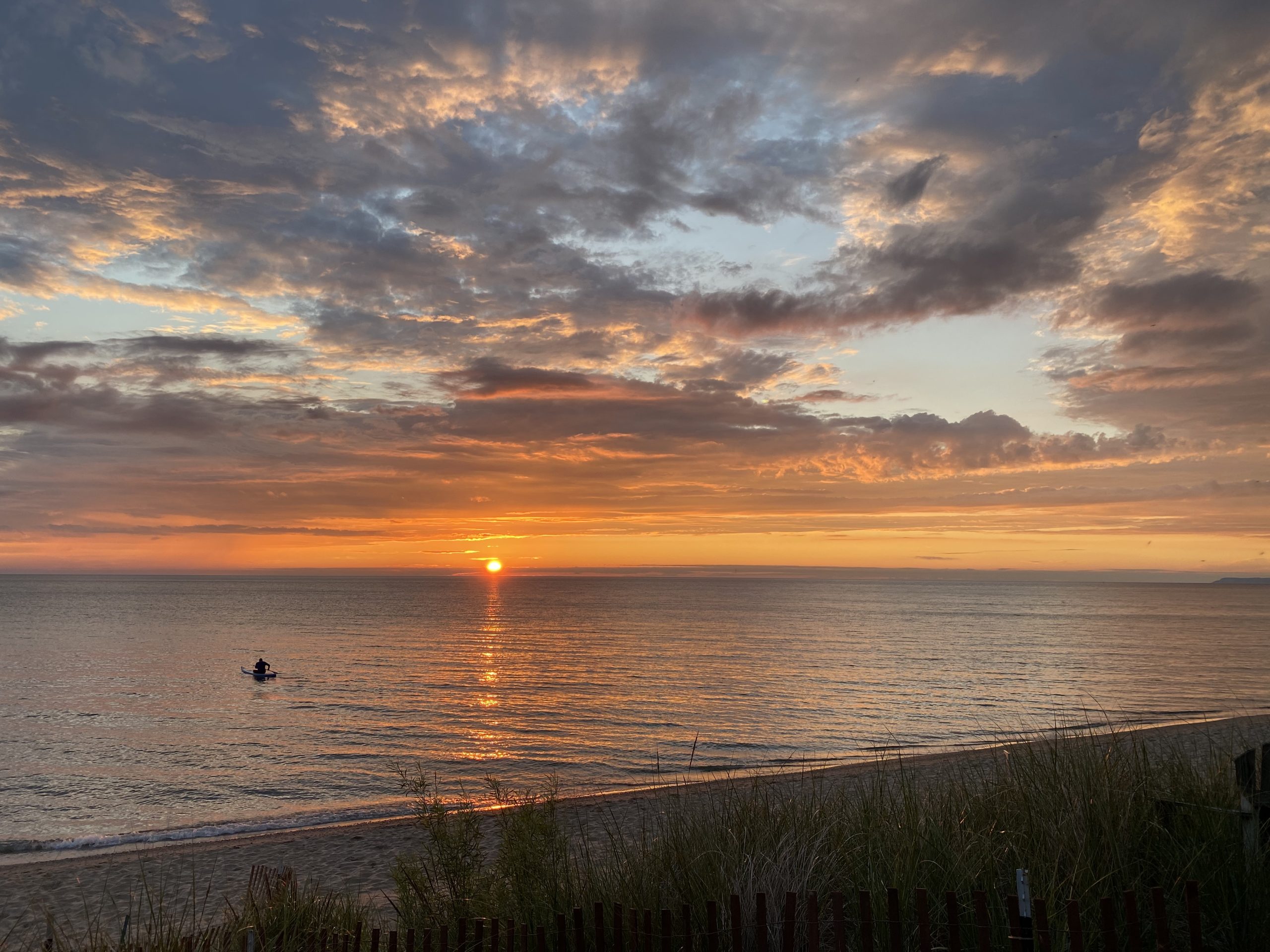 Sun setting beneath layered clouds over the lake with beach grass in the foreground