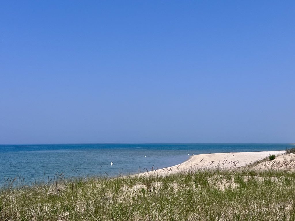 Pale sandy shoreline with beach grass and clear blue water under a hazy sky