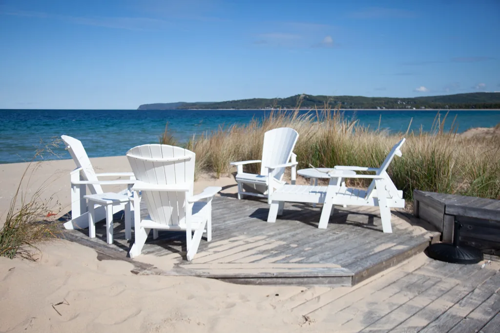 White Adirondack chairs and a small table on a sandy deck with beach grass and blue water beyond